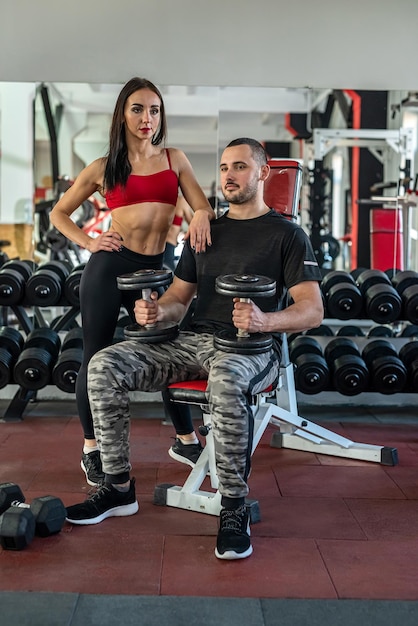 Entrenador masculino ayuda a su cliente mujer a trabajar con pesas con mancuernas en el gimnasio
