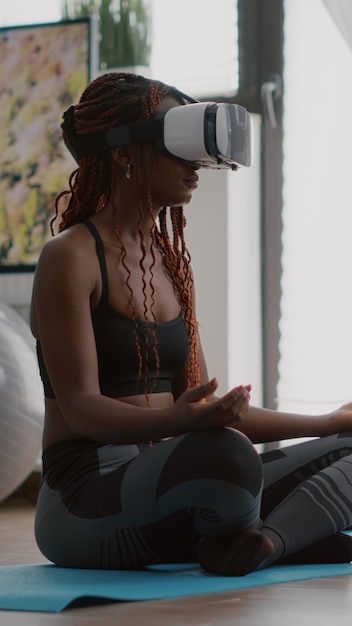 Entrenador negro mujer con casco de realidad virtual mientras está sentado en el mapa de yoga