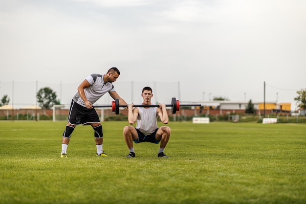 Entrenador personal ayudando a su aprendiz a levantar pesas mientras está de pie en el campo de fútbol.