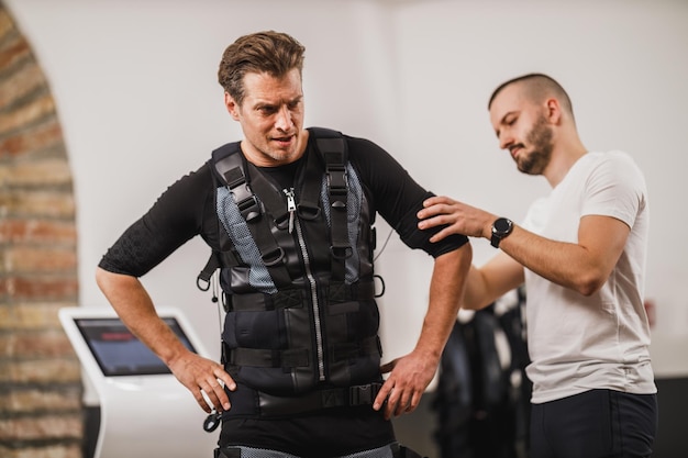 El entrenador personal está ayudando a un hombre de mediana edad a vestirse para EMS trabajando en el gimnasio.