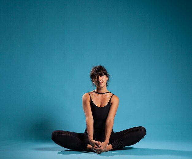 Entrenador personal sentado en posición de loto en una alfombra de yoga estirando los músculos del cuerpo en un estudio con fondo azul. Mujer atleta con ropa deportiva practicando la postura de padmasana para un estilo de vida saludable