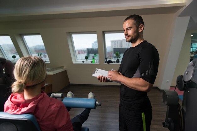 Entrenador personal toma notas mientras la mujer joven ejercita las piernas en la máquina en el gimnasio