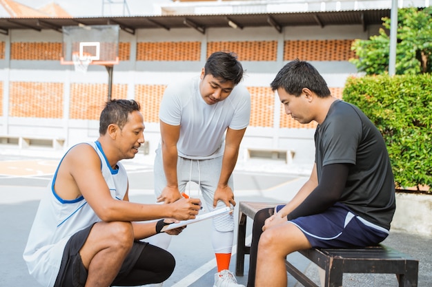 Entrenador sosteniendo un portapapeles dando instrucciones a dos jugadores de baloncesto