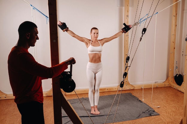 El entrenador tira de las cuerdas estirando a una chica con uniforme deportivo en un simulador para estirar el cuerpo humano