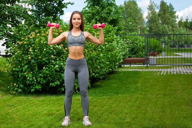 Una entrenadora de cabello oscuro con un top corto deportivo y mallas de gimnasia hace un swing con la mano en los lados con mancuernas en un día de verano en un parque en un césped verde