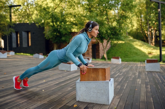 Entrenamiento de ajuste matutino sobre colchoneta en el parque, mujer en auriculares haciendo ejercicios de flexión de brazos