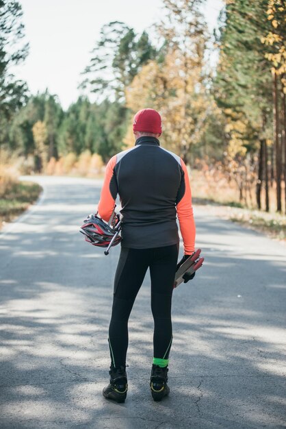 Entrenamiento de un atleta en los patinadores Biatlón paseo en los patines con bastones de esquí en el casco Entrenamiento de otoño Deporte de rodillos El atleta va y sostiene equipo deportivo en su mano