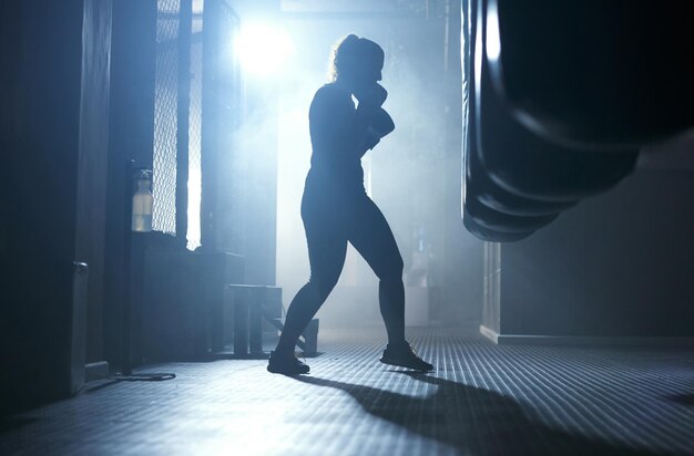 Entrenamiento de boxeo y persona en el gimnasio oscuro y práctica con saco de boxear acción y energía para el deporte Ejercicio saludable y boxeador con habilidades para la competencia entrenamiento y rendimiento de luchador