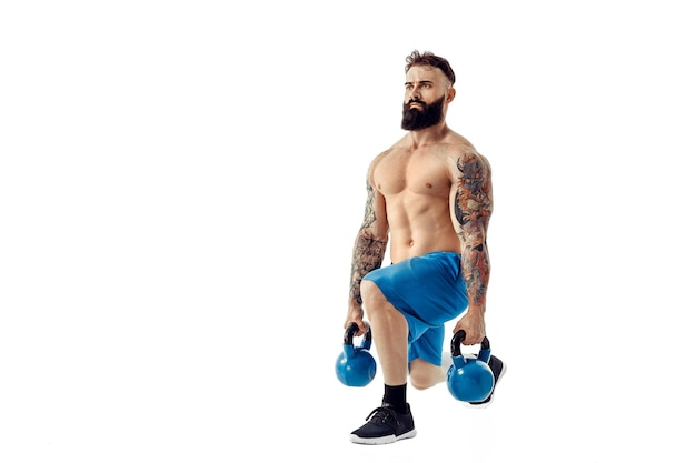 Entrenamiento de culturista de atleta masculino barbudo tatuado sin camisa muscular con pesas rusas sobre un fondo blanco Aislar