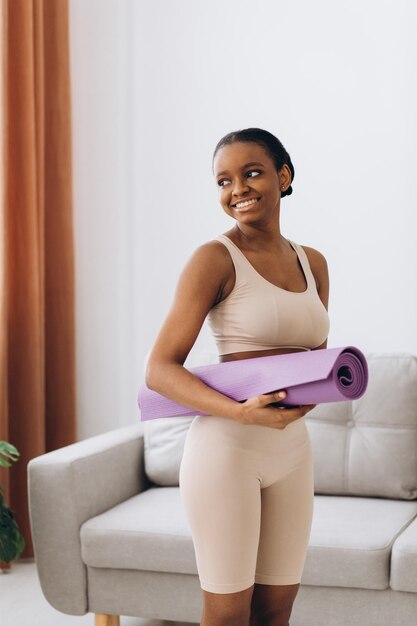 Entrenamiento doméstico Joven mujer negra deportiva sosteniendo una estera de yoga en casa entrenando en la sala de estar