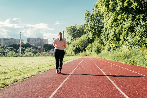 Entrenamiento en el estadio, al aire libre. Mujer joven de talla grande caminando afuera en un día soleado de verano. Mujer con sobrepeso haciendo deporte, vista posterior. Estilo de vida activo, pérdida de peso.