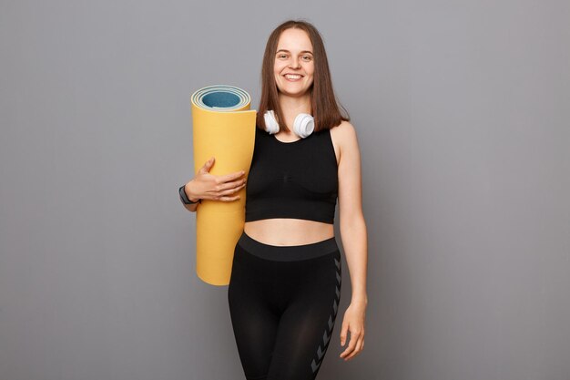 Entrenamiento estilo de vida de yoga Joven hermosa mujer caucásica con ropa deportiva sosteniendo estera de yoga aislada en fondo gris preparándose para hacer ejercicio en el gimnasio