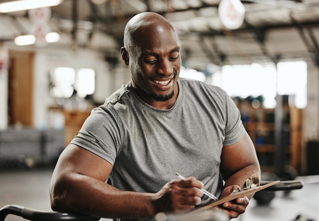 Entrenamiento de gimnasio y entrenador personal con lista de verificación en consultoría de portapapeles para entrenamiento deportivo en gimnasio Hombre negro de entrenador de fitness musculoso activo y sonriente escribiendo sobre salud, bienestar y ejercicio