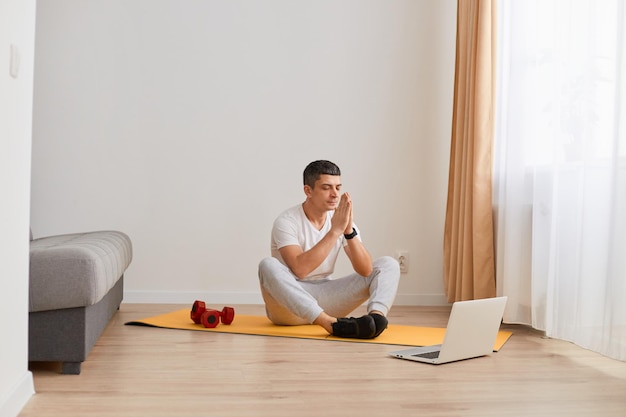 Entrenamiento en línea con aparatos Toma horizontal de un tipo con reloj inteligente haciendo yoga viendo videos de entrenamiento en casa en la sala de estar durante el día mantiene las palmas en gesto de oración