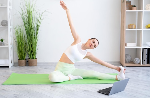 Entrenamiento en línea Fitness doméstico Deporte en interiores Mujer atlética motivada practicando ejercicio físico de estiramiento en colchoneta de yoga en el tutorial de entrenamiento de portátiles en casa