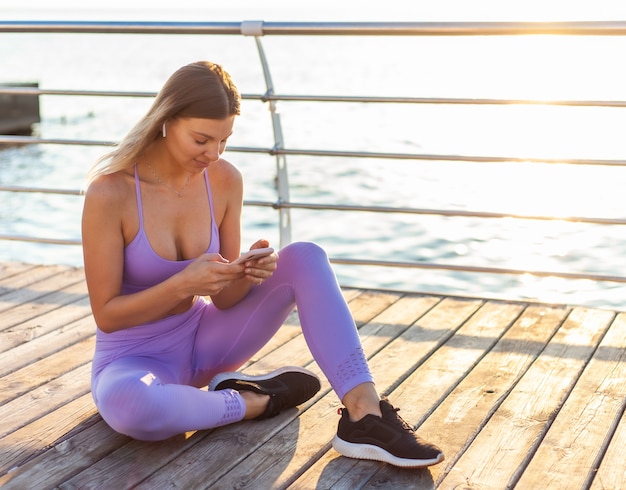 Entrenamiento de la mañana. Joven mujer deportiva en ropa deportiva utiliza teléfono inteligente mientras está sentado en la playa al amanecer