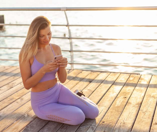 Entrenamiento de la mañana. Joven mujer deportiva en ropa deportiva utiliza teléfono inteligente mientras está sentado en la playa al amanecer