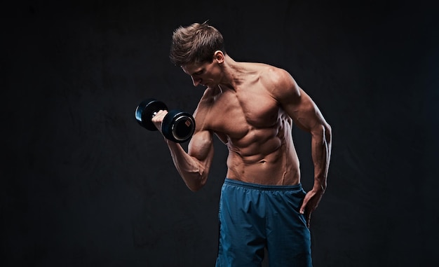 Entrenamiento con mancuernas de bíceps masculino sin camisa atlético sobre fondo gris oscuro.