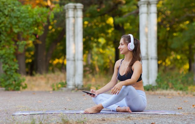Entrenamiento y recreación al aire libre. Mujer joven deporte escuchando música con auriculares mientras está sentado en la estera de yoga en el parque en un día soleado