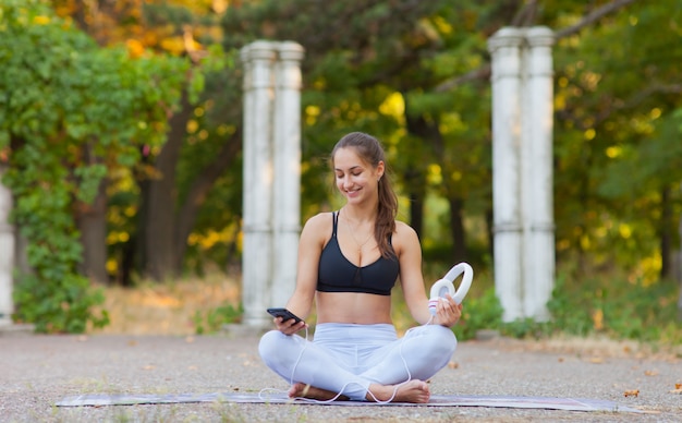 Entrenamiento y relajación al aire libre. Mujer joven en ropa deportiva descansando ejercicios de yoga previos a la práctica Ñ † Ñ € ÑˆÐ´Ñƒ sosteniendo los auriculares en la mano con un teléfono inteligente