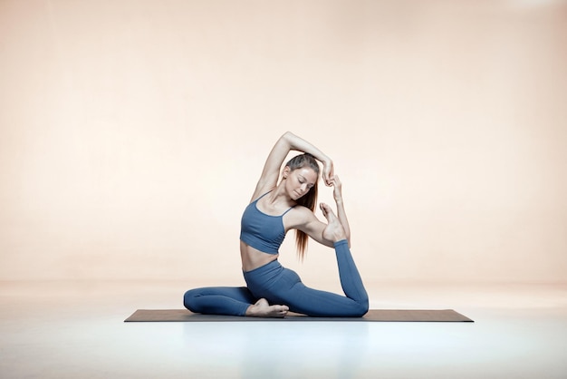 Entrenamiento de yoga o pilates Chica joven flexible estirar los músculos del cuerpo haciendo King Pigeon Pose en estudio sobre fondo beige Foto de alta calidad