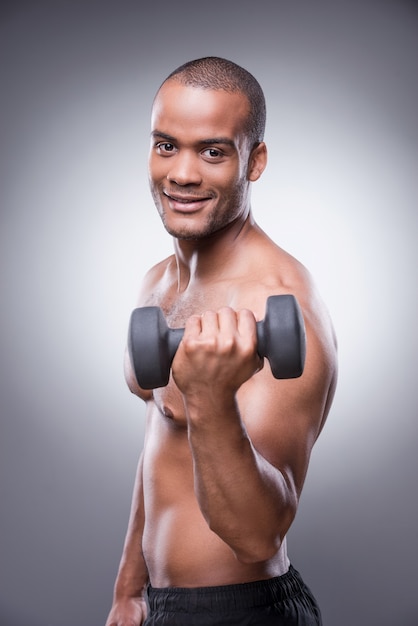 Entrenando su cuerpo a la perfección. Joven africano sin camisa haciendo ejercicio con mancuernas y sonriendo