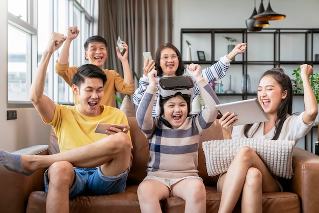 Entretener el estilo de vida familiar de la tecnología, adicto a la familia de múltiples generaciones, a los lentes vr de teléfonos inteligentes y dispositivos de tableta, todos jugando en línea con diversión y alegría, saludando la victoria juntos en casa