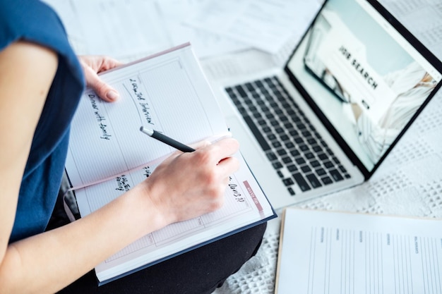 Entrevista de trabajo Encontrar nuevo trabajo Mujer joven escribiendo en el diario de Planner notas sobre una entrevista de trabajo sentada cerca de una computadora portátil en la cama en casa