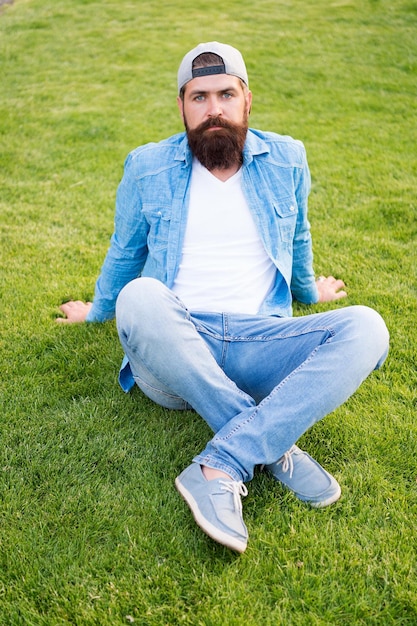 Entscheiden Sie sich für Einfachheit statt Auffälligkeit. Modemann, der auf grünem Gras sitzt. Bärtiger Mann im modischen Denim-Stil im Freien. Trendiger Hipster mit langem Bart und modischer Mütze nach hinten. Herren Sommermode.