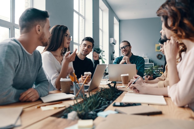 Entschlossen, der Beste zu sein. Gruppe junger moderner Menschen in eleganter Freizeitkleidung, die etwas diskutieren und lächeln, während sie im Kreativbüro arbeiten working