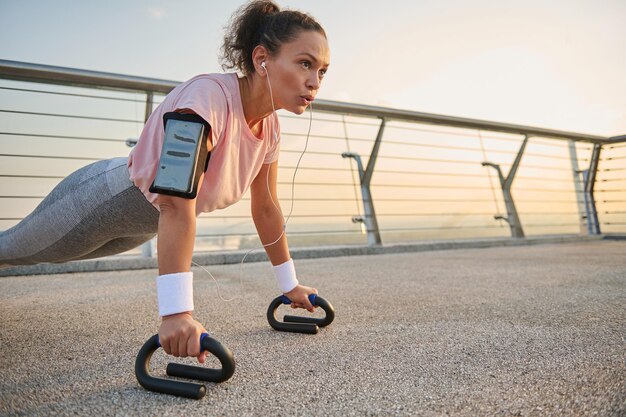 Entschlossene Sportlerin, afroamerikanische Sportlerin mit Kopfhörern und Smartphone-Halterung beim Trainieren, Liegestützen, Liegestützen, während sie bei Sonnenaufgang ein Körpergewichtstraining auf der Stadtbrücke durchführt