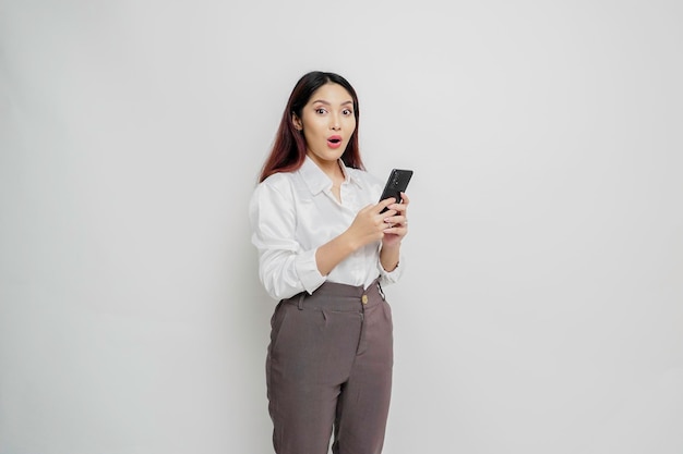 Entsetzte asiatische Frau, die weißes Hemd trägt und ihr Telefon lokalisiert durch weißen Hintergrund hält