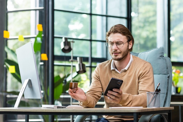 Entsetzter Geschäftsmann, der im Büro mit einem Computer sitzt und eine Kreditkarte in seiner Hand hält, er