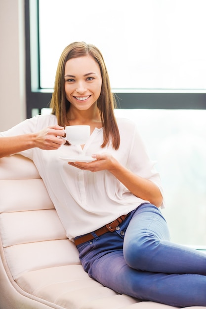 Entspannen bei einer Tasse frischem Kaffee. Schöne junge Frau, die Kaffeetasse hält und lächelt, während sie am Stuhl sitzt