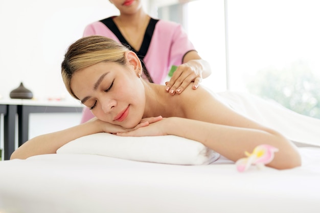 Entspannende junge asiatische Frau, die sich auf Massagebetten hinlegt und die Augen schließt, während sie eine Massage von einem Therapeuten in einem asiatischen Luxus-Spa-Salon und einem Wellness-Center erhält und auf das Spa-Salon-Konzept wartet