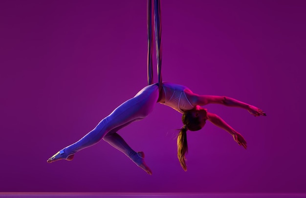 Entspannendes junges flexibles Mädchen, das Luftfliegen-Yoga-Training über violettem Studiohintergrund in Neon macht