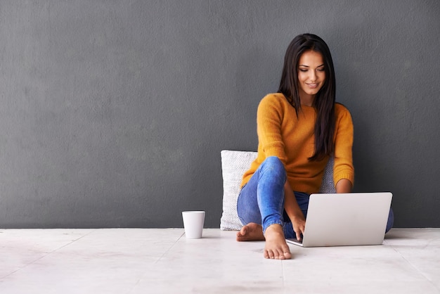 Entspannt und glücklich auf dem Boden Aufnahme einer attraktiven jungen Frau, die einen Laptop benutzt, während sie auf dem Boden sitzt