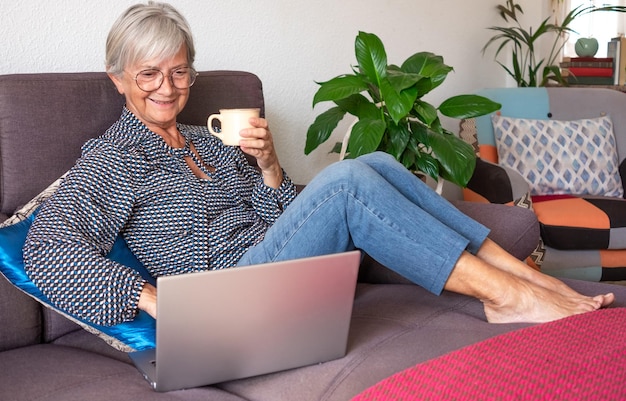 Entspannte ältere Frau, die zu Hause am Laptop arbeitet und auf dem Sofa im Wohnzimmer sitzt Ältere Dame, die eine Tasse Kaffee hält und Technik und Soziales genießt