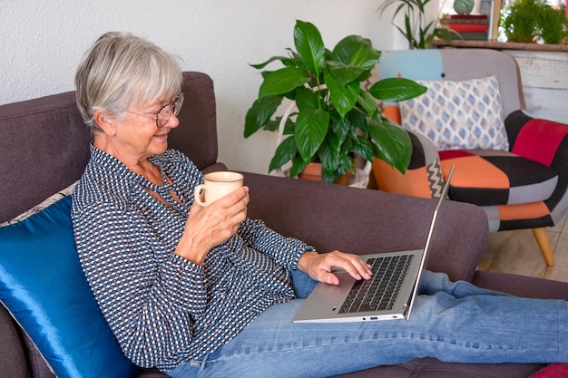 Entspannte ältere Frau, die zu Hause auf dem Laptop surft und auf dem Sofa im Wohnzimmer sitzt Moderne ältere Dame, die eine Kaffeetasse hält