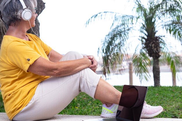 Entspannte ältere frau mit kopfhörern, die draußen sitzen und auf das meer schauen. laptop-computer auf werkbank