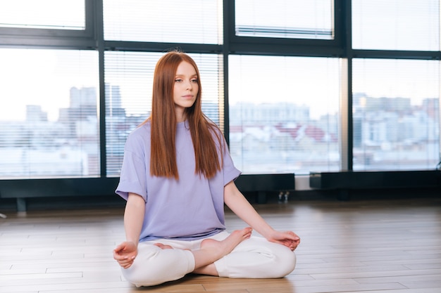 Entspannte, attraktive junge Frau, die auf dem Boden im Lotussitz auf dem Hintergrund des Fensters im hellen Büroraum meditiert.