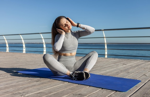 Entspannte Fitnessfrau, die Musik mit Kopfhörern hört, während sie auf einer Matte am Strand sitzt Sport und Entspannung