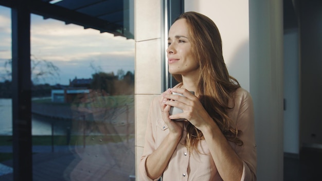 Entspannte Frau, die nach dem Arbeitstag am Fenster bleibt. Hübsche Dame, die den Sonnenuntergang beobachtet