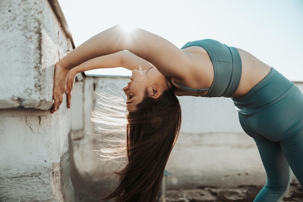 Entspannte Frau, die Yoga-Dehnübungen in der Nähe einer Wand auf einem Dach bei Sonnenuntergang praktiziert.