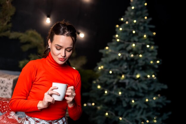 Entspannte Frau im roten Pullover posiert mit einer Tasse heißem Getränk in der Nähe des Neujahrsbaums Stilvolle Dekorationen vor der Festzeit