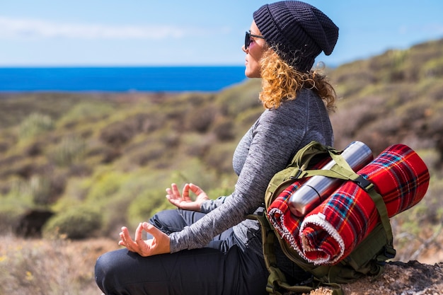 Entspannte Frau in Meditationsposition, die auf den Felsen während der Freizeitaktivität des Reiserucksackabenteuers im Freien sitzt. Erwachsene Frauen genießen aktiven Lebensstil und Natur