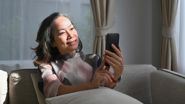 Entspannte Frau mittleren Alters, die mit dem Handy soziale Medien überprüft oder Online-Nachrichten auf einem bequemen Sofa zu Hause liest Technologiekonzept für den Ruhestand