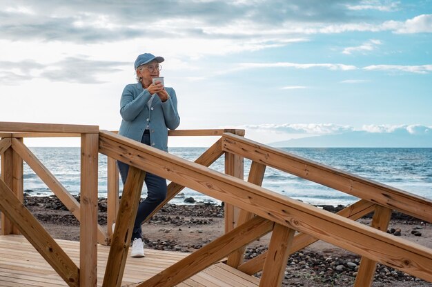 Entspannte, gutaussehende Seniorin mit Mütze und Brille, die bei Sonnenaufgang im Freien steht und eine Tasse Kaffee oder Tee hält Lächelnde ältere Dame, die die Stille des Morgens am Strand genießt