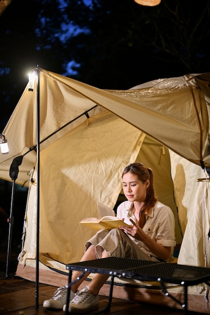 Entspannte junge Asiatin sitzt vor ihrem Campingzelt und liest nachts ein Buch