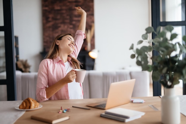 Entspannte junge Dame, die eine Kaffeetasse hält und ihren Arm nach oben streckt, während sie am Schreibtisch sitzt